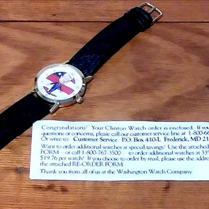 May include: A wristwatch with a black leather band and a gold-colored case. The watch face features a design with red, white, and blue colors. The watch is on a wooden surface.