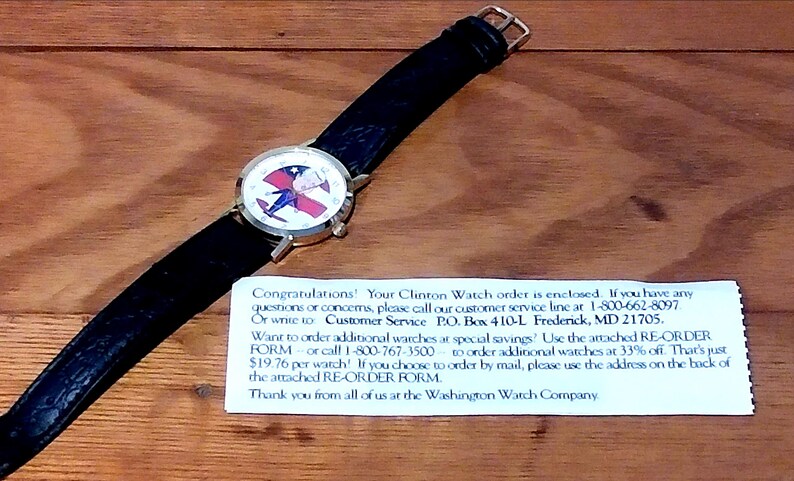 May include: A wristwatch with a black leather band and a gold-colored case. The watch face features a design with red, white, and blue colors. The watch is on a wooden surface.