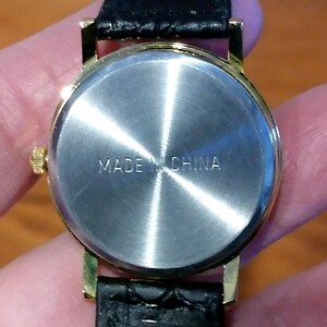 May include: Close-up of the back of a gold-toned wristwatch with a black leather band. The watch face is silver with the words "MADE IN CHINA" stamped on it. The watch is held by a person.