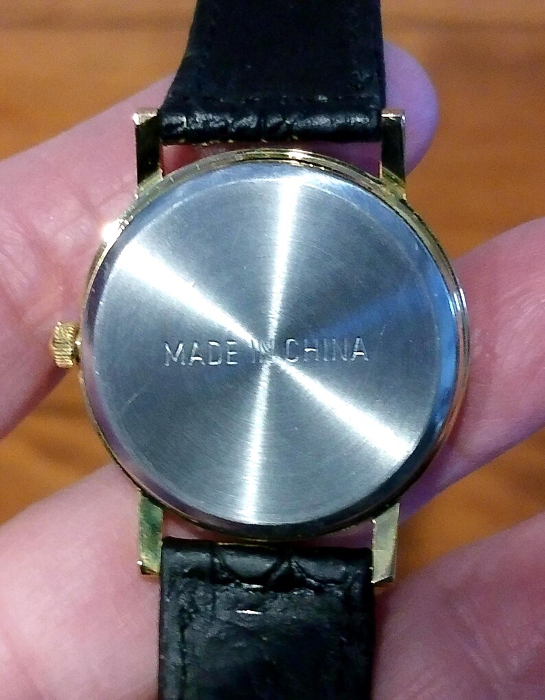 May include: Close-up of the back of a gold-toned wristwatch with a black leather band. The watch face is silver with the words "MADE IN CHINA" stamped on it. The watch is held by a person.