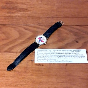 May include: A wristwatch with a black leather band and a round face featuring a patriotic design. The watch is set against a wooden surface. The watch face has a red, white, and blue design.