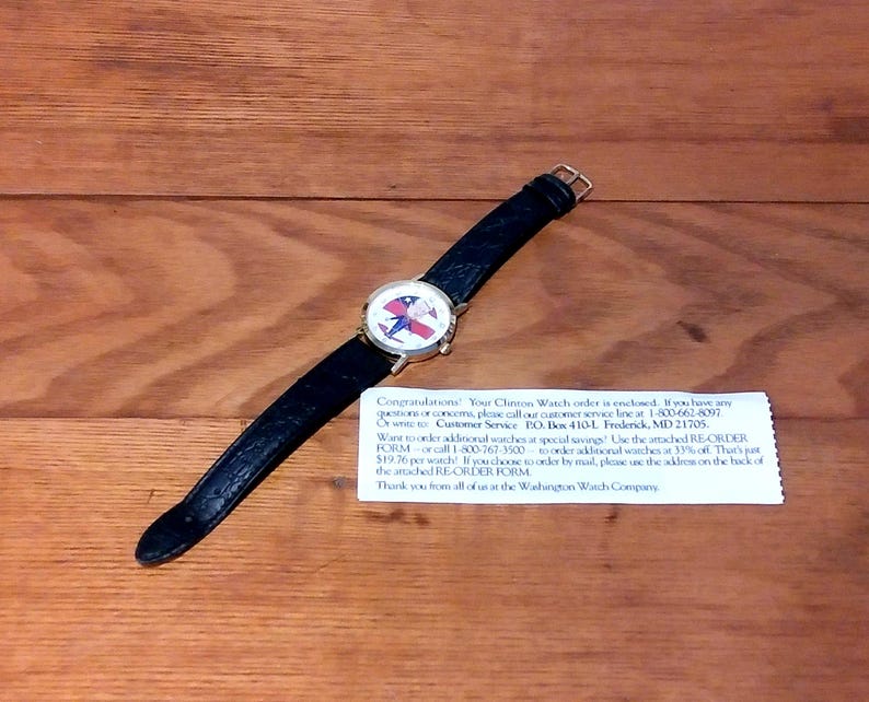 May include: A wristwatch with a black leather band and a round face featuring a patriotic design. The watch is set against a wooden surface. The watch face has a red, white, and blue design.