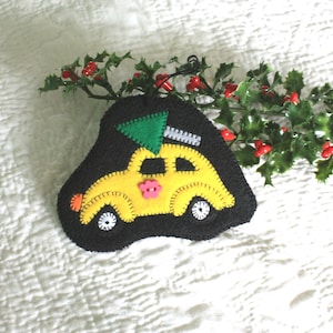 May include: A handmade Christmas ornament featuring a yellow car with a green tree on top, set against a black background. The ornament has white wheels and a pink flower. Holly with red berries is in the background.
