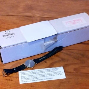 May include: A wristwatch with a black leather band and a silver-colored face rests on a wooden surface next to a white box. The watch band is labeled "GENUINE LEATHER". A small white card with text is also present.