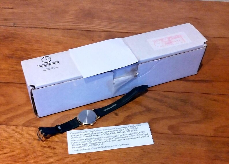 May include: A wristwatch with a black leather band and a silver-colored face rests on a wooden surface next to a white box. The watch band is labeled "GENUINE LEATHER". A small white card with text is also present.