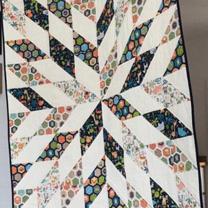 Könnte beinhalten: Eine handgefertigte Steppdecke mit Sternmuster. Die Steppdecke hat weiße, marineblaue, orangefarbene, grüne und beige geometrische Muster. Die Steppdecke hängt an einer Stange mit Clips.