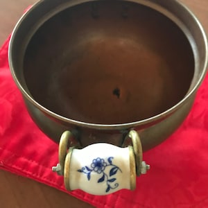 May include: A copper serving bowl with two porcelain handles decorated with blue floral designs. The bowl has a shiny, metallic finish and is sitting on a red surface.