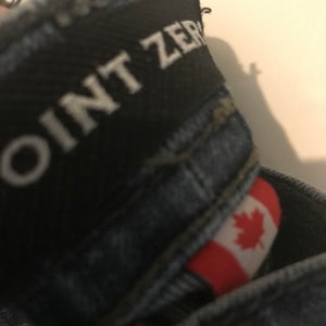 Peut inclure: Gros plan sur une paire de jeans en denim bleu avec une étiquette noire sur laquelle est inscrit "JOINT ZERO" et un petit écusson du drapeau canadien.