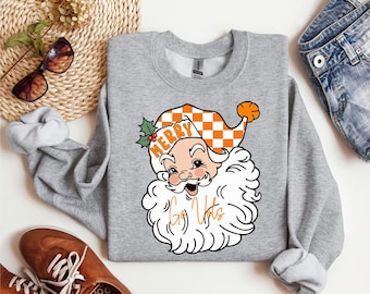 May include: A light gray sweatshirt featuring a cartoon Santa Claus with a checkered orange and white hat. The Santa graphic includes the words "Merry" and "Go Vols" in orange and white. The sweatshirt has long sleeves.
