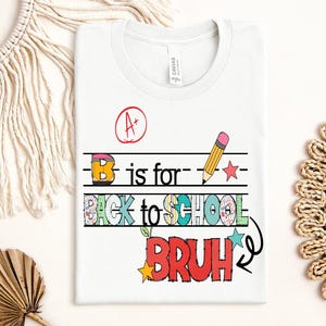 Può includere: T-shirt bianca con un design grafico colorato. Il design include il testo "B is for BACK TO SCHOOL BRUH" con una matita, stelle e un simbolo di voto A+. La maglietta è realizzata in materiale morbido.