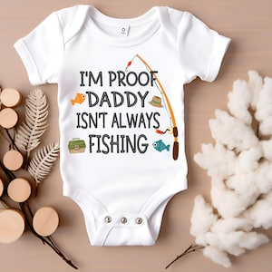 Puede incluir: Un body blanco para bebé con un gráfico negro que dice "Soy la prueba de que papá no siempre está pescando". El gráfico incluye una caña de pescar, un pez, un sombrero de pesca y una caja de aparejos.