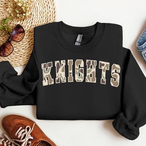 May include: Black crewneck sweatshirt with the word "KNIGHTS" in a camouflage pattern across the chest. The sweatshirt is surrounded by accessories, including sunglasses, a woven mat, and a pair of brown shoes.