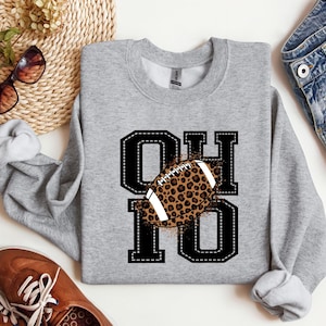 Könnte beinhalten: Ein meliertes graues Sweatshirt mit den Buchstaben "OHIO" in Schwarz, umrandet von weißen Nähten. Ein Football mit Leopardenmuster befindet sich in der Mitte der Buchstaben. Das Sweatshirt ist ein lässiger, bequemer Stil.