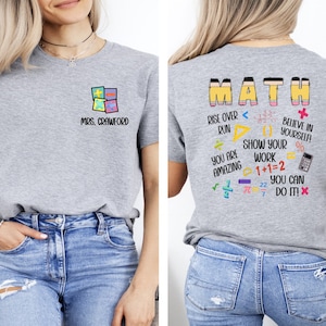 May include: Gray t-shirt with a math-themed design on the back. The design includes the word "MATH" in large letters, along with various math symbols, equations, and motivational phrases such as "Rise Over Run" and "You Can Do It!" The front of the shirt has a small design of colorful math symbols and the text "Mrs. Crawford".
