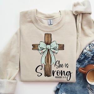 May include: A beige sweatshirt featuring a graphic of a wooden cross with a light blue bow and the text "She is Strong Proverbs 31:25".
