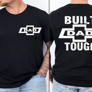 May include: Black t-shirt with a white graphic on the front that says "DAD" inside a stylized emblem. The back of the shirt features the words "BUILT DAD TOUGH" in white block letters.