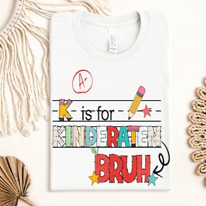 May include: White t-shirt with a colorful graphic that reads "K is for KINDERATEN BRUH". The design includes a pencil, stars, and an A+ grade symbol. The shirt is made of a soft material.