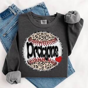 Peut inclure: Un sweat-shirt gris foncé avec un motif de baseball portant le mot "Dragons" en blanc. Le baseball a un centre à imprimé léopard, des coutures rouges et un petit cœur rouge. Le sweat-shirt est présenté sur un jean bleu.