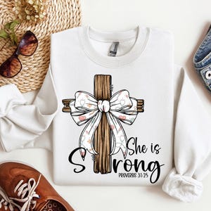 Può includere: Felpa bianca con una croce di legno marrone decorata con un fiocco bianco e rosa. Il testo "She is Strong" e "Proverbs 31:25" è stampato sotto la croce.