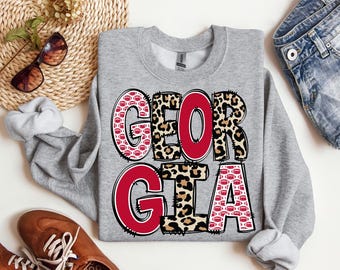 Op de afbeelding: Een grijze sweatshirt met het woord "GEORGIA" in grote, vetgedrukte letters. De letters hebben een mix van patronen, waaronder rode voetballen, luipaardprint en effen rood. De sweatshirt ligt plat met een zonnebril, een gevlochten mand en een spijkerbroek.