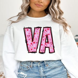 May include: White sweatshirt with a pink and red "VA" graphic featuring a heart pattern. The sweatshirt has long sleeves and a crew neck.