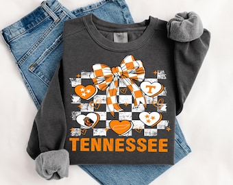 Może przedstawiać: Ciemnoszara bluza z motywem Tennessee. Projekt zawiera wzór w szachownicę, pomarańczową kokardę, kształty serc i słowo "Tennessee" w kolorze pomarańczowym. Bluza jest prezentowana na niebieskich dżinsach.