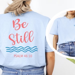 May include: Light blue t-shirt with the words "Be Still" in pink script on the back, along with blue wave graphics and "Psalm 46:10." A smaller wave graphic is on the front. The shirt is a casual wear item.