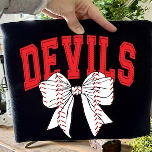 Puede incluir: Bolso tote negro con la palabra "DEVILS" en letras rojas. Un lazo blanco con costuras de béisbol rojas está centrado debajo. El bolso lo sostiene una persona.