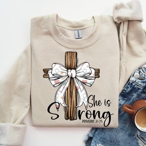 May include: Beige sweatshirt with a wooden cross design and a white bow decorated with hearts and floral patterns. The text "She is Strong" and "Proverbs 31:25" are printed in black. A coffee mug and denim jeans are in the background.