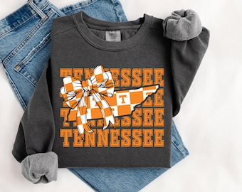 Może przedstawiać: Ciemnoszara bluza z konturem stanu Tennessee w pomarańczowo-białą kratę, z pasującą kokardą. Słowo "Tennessee" powtarza się w kolorze pomarańczowym. Widoczne podwinięte rękawy i niebieskie dżinsy.