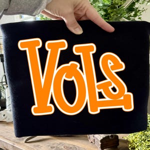Puede incluir: Una camiseta negra con la palabra VOLS impresa en letras grandes, negritas y de color naranja con un borde blanco. El texto está centrado en la parte delantera de la prenda, que está confeccionada con una tela negra sólida y uniforme.
