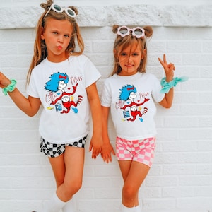 Könnte beinhalten: Zwei junge Mädchen tragen weiße T-Shirts mit einer bunten Grafik einer Figur mit blauen Haaren und dem Text "Little Miss". Ein Mädchen trägt schwarz-weiß karierte Shorts, das andere rosa karierte Shorts.