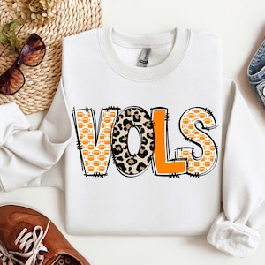 Puede incluir: Sudadera blanca con la palabra "VOLS" en letras grandes y en negrita. Las letras están decoradas con patrones de fútbol americano, estampado de leopardo y naranja liso. La sudadera tiene mangas largas.
