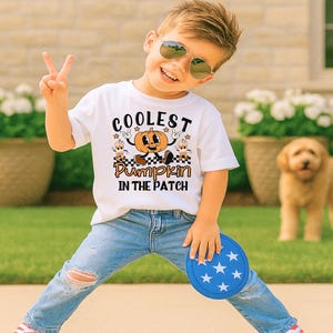Può includere: T-shirt bianca con la scritta "Coolest Pumpkin in the Patch" e una grafica di zucca dei cartoni animati. La maglietta è abbinata a jeans blu e un frisbee blu. L'immagine è di un bambino con occhiali da sole.