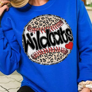 May include: A royal blue sweatshirt featuring a glittery baseball graphic. The baseball has a leopard print design and the word "Wildcats" written in black across it. A small red heart is on the side of the baseball.