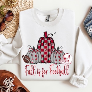 Op de afbeelding: Witte sweatshirt met een afbeelding van drie pompoenen, een voetbal en een helm. De centrale pompoen heeft een rood-wit geruit patroon. De tekst "Fall is for Football" staat in rode letters.