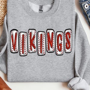 Puede incluir: Sudadera gris con la palabra "VIKINGS" escrita en letras blancas con costuras rojas, que se asemejan a pelotas de béisbol.