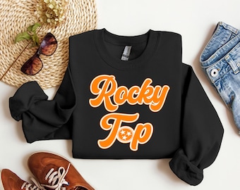 Puede incluir: Sudadera negra con texto naranja que dice "Rocky Top" con un contorno blanco. El texto incluye un pequeño círculo naranja con tres estrellas blancas en el centro.