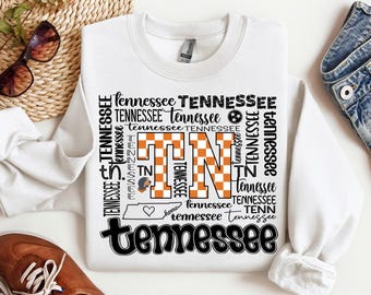 May include: White sweatshirt with a black and orange graphic design featuring the letters "TN" in a checkered pattern and the word "Tennessee" repeated multiple times. The graphic also includes a small heart and a state outline.