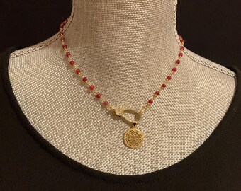 Pode incluir: Um colar de corrente dourada com pequenas contas redondas de cor vermelha translúcida espaçadas uniformemente. O colar inclui um fecho lagosta dourado adornado com pequenas pedras claras e um pingente circular dourado com padrão.