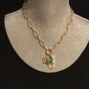 May include: A gold chain necklace with a green gemstone pendant and a charm. The pendant is shaped like a leaf and has a small charm attached to it.