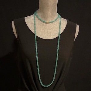 May include: Two necklaces on a mannequin. The top necklace is a choker style with small, light blue beads and a gold-colored bead. The second necklace is long, with similar light blue beads. The mannequin is wearing a black top.