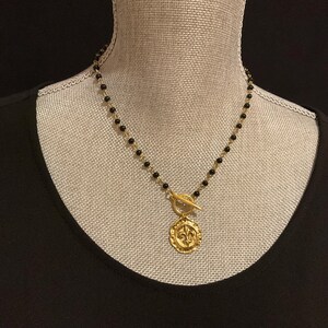 May include: Gold-tone necklace with a black bead chain and a fleur-de-lis pendant. The pendant is round with a textured surface and a toggle clasp. The necklace is displayed on a mannequin.