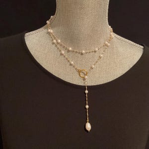 May include: A layered necklace featuring delicate strands of white pearls and gold-colored chain. The necklace has a circular pendant and a longer strand of pearls that dangles down. The necklace is displayed on a mannequin.