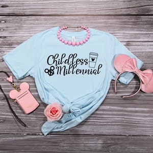 May include: Light blue t-shirt with the text "Childless Millennial" and a coffee cup with a Mickey Mouse head on it. The shirt is on a wooden surface with a pink headband, a pink purse with Mickey Mouse ears, and a pink rose.