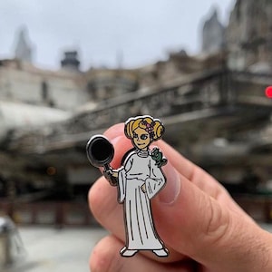 以下が含まれることがあります： スターウォーズのレイア姫の漫画キャラクターがフライパンを持っている銀色のエナメルピン。キャラクターは白いドレスを着ており、頭に花冠をかぶっています。ピンは街のぼやけた背景に置かれています。