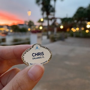 May include: A gold name tag with a white background and black lettering. The name tag says "CHRIS ORLANDO, FL".