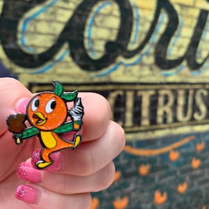 May include: Orange Bird enamel pin with a green leaf, holding a brown ice cream cone and a white ice cream cone. The pin is in front of a yellow brick wall with the word "Citrus" written in black.