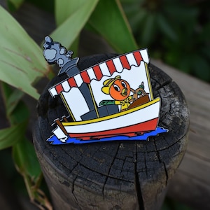 May include: A colorful enamel pin featuring a cartoon orange bird driving a small boat with a red and white striped awning. The boat has a black smokestack and a white flag.