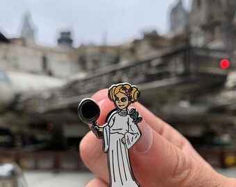 Peut inclure: Une épingle en émail argentée représentant un personnage de dessin animé de la princesse Leia tenant une poêle. Le personnage porte une robe blanche et une couronne de fleurs sur la tête. L'épingle est placée sur un fond flou d'une ville.